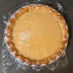 a photo of a butterscotch pie filled and ready to cool with plastic wrap over the top to prevent a skin from forming on the custard