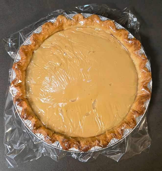 a photo of a butterscotch pie filled and ready to cool with plastic wrap over the top to prevent a skin from forming on the custard