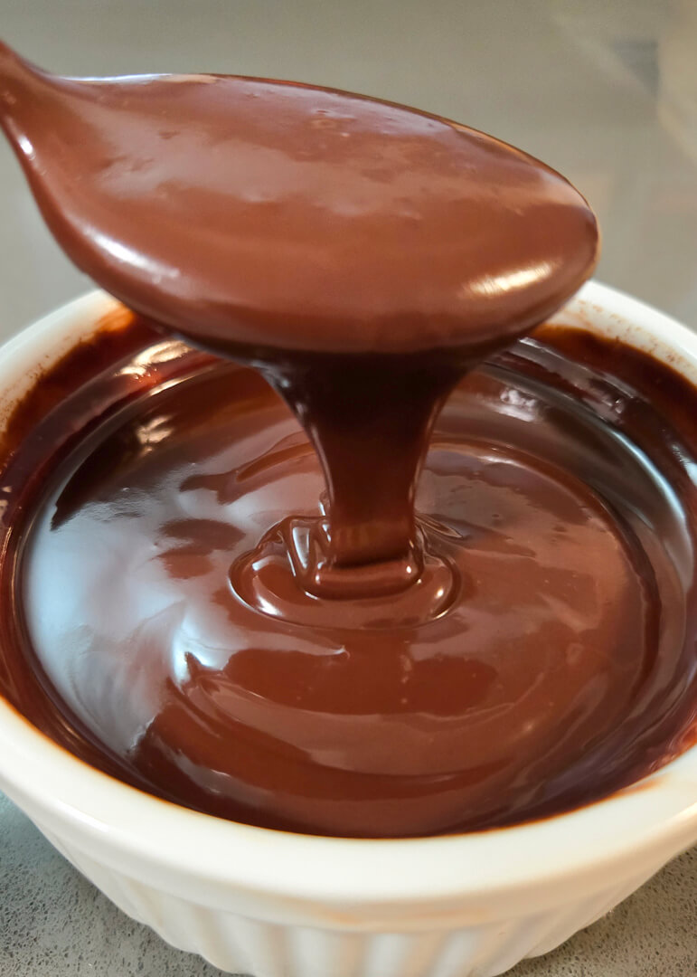 close up photo of chocolate ganache recipe coming off of a spoon and falling into a bowl in smooth ribbons