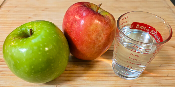 a green granny smith apple, a red gala apple, and a glass of water on a cutting board