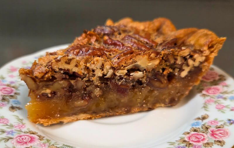 close up shot of a slice of pecan pie with a glossy filling, candied pecans, and buttery crust