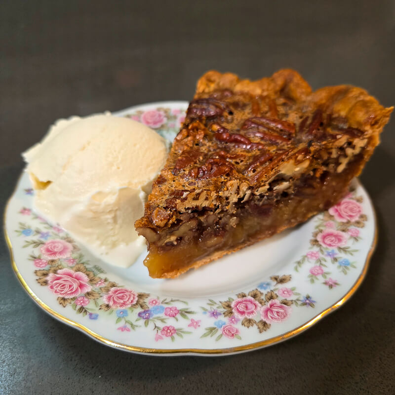 a slice of pecan pie with glossy filling and candied pecans on top with a golden buttery crust and a scoop of vanilla ice cream on a floral plate