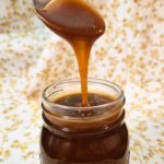 a photo on a festive background of a mason jar with caramel sauce and spoon above it pouring caramel sauce down into the jar for Salted Caramel Sauce Recipe
