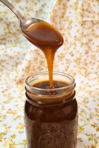 a photo on a festive background of a mason jar with caramel sauce and spoon above it pouring caramel sauce down into the jar for Salted Caramel Sauce Recipe
