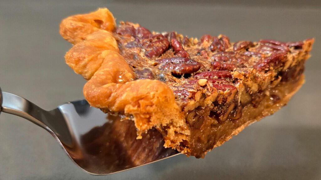 a close up photo of a slice of bourbon pecan pie with glossy caramel filling and candied pecans in a buttery crust