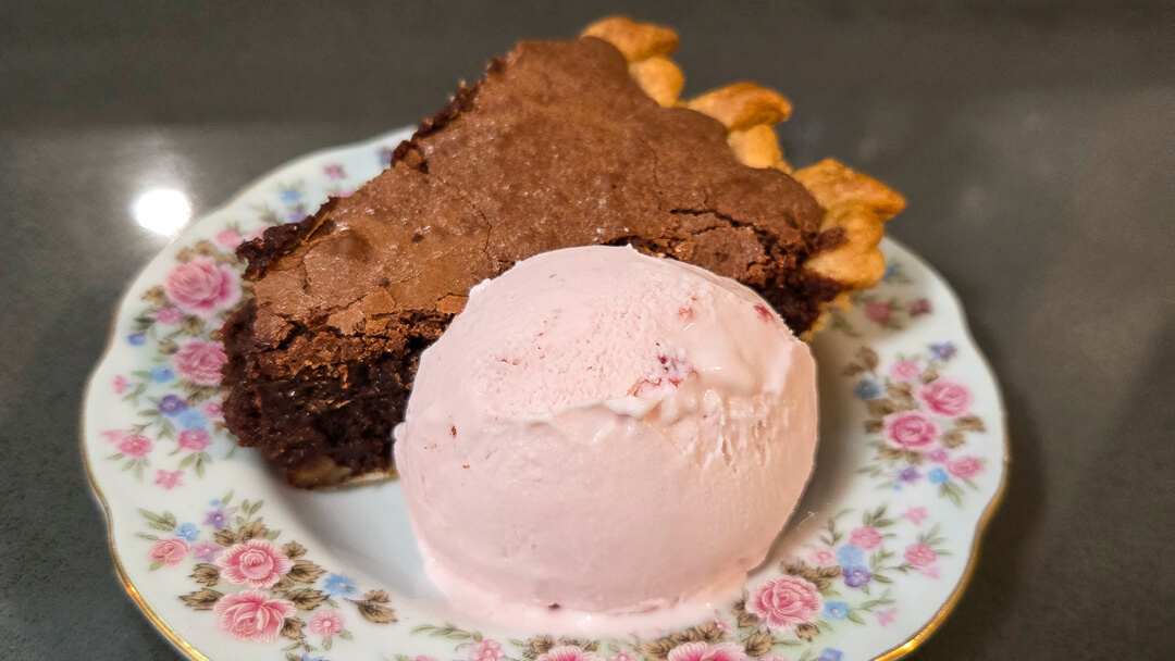 Slice of rich brownie pie served with a scoop of strawberry ice cream on a floral dessert plate.