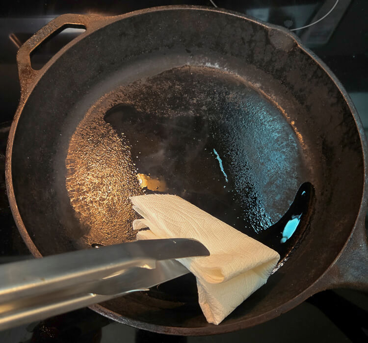 seasoning a cast iron pan by spreading oil with a paper towel and tongs