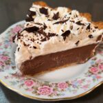 a slice of chocolate cream pie on a floral plate showing whipped cream with chocolate shavings, a flaky pastry crust, and rich chocolate pudding