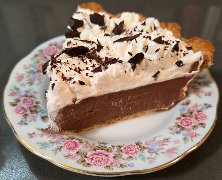 a slice of chocolate cream pie on a floral plate showing whipped cream with chocolate shavings, a flaky pastry crust, and rich chocolate pudding