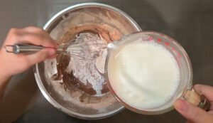 process photo showing how to temper eggs in chocolate pudding. Show chocolate pudding being whisked vigorously as a small amount of milk is poured in