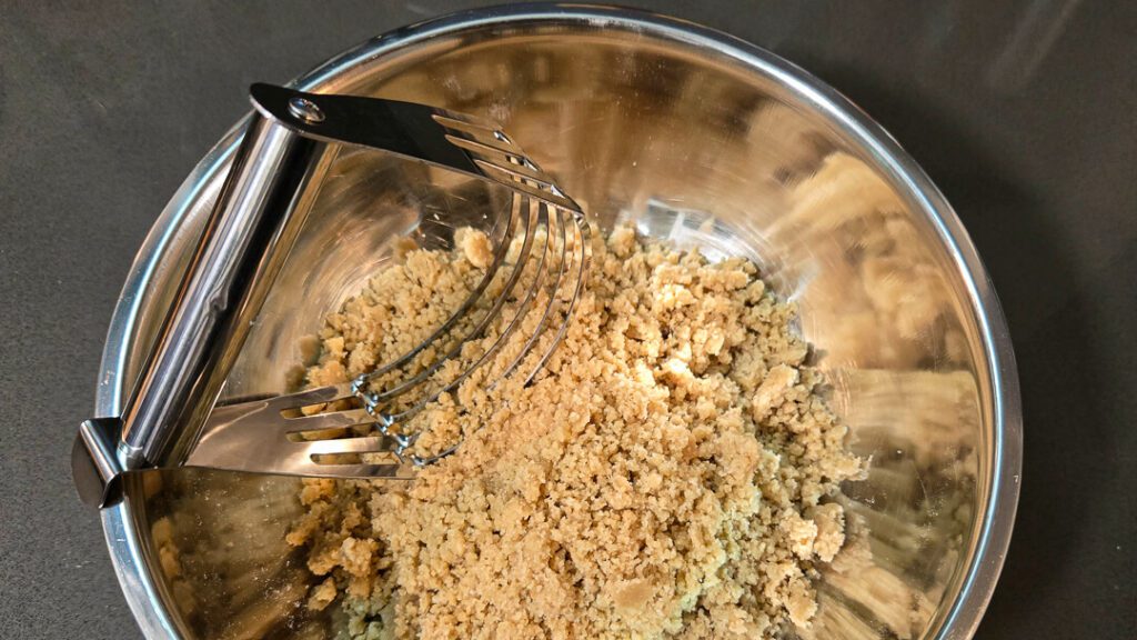 Close-up of crumble topping being mixed in a metal bowl with a pastry cutter, showing coarse buttery crumbs for pie topping.