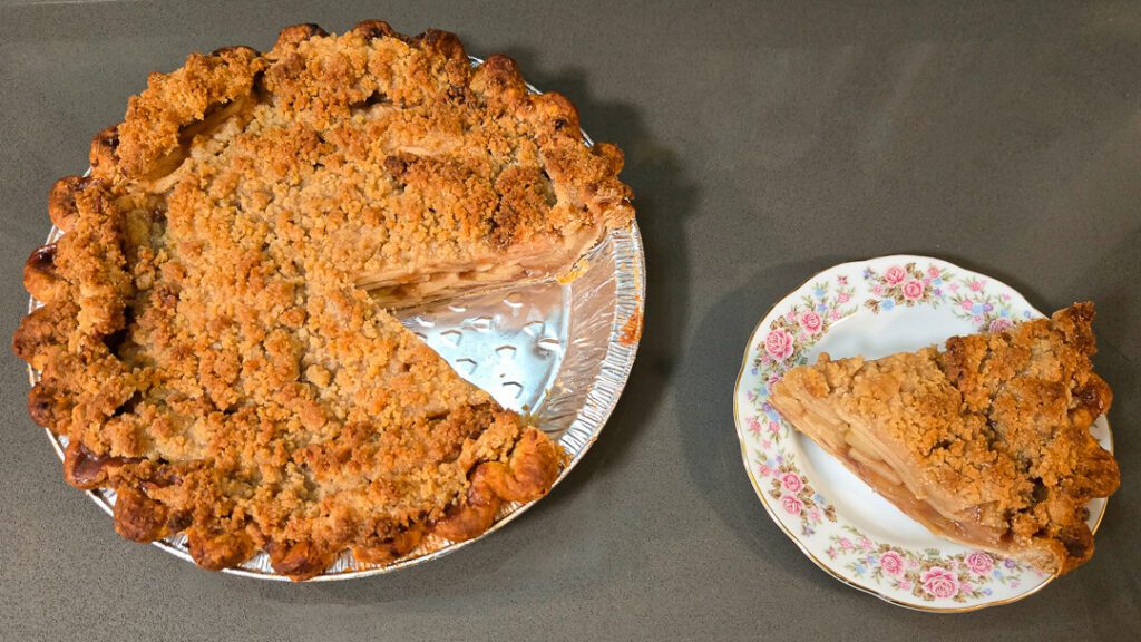 Overhead view of a Dutch apple pie with a golden crumble topping, one slice removed and served on a floral plate beside the pie.