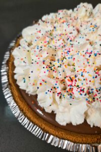Close up photo of a chocolate cream pie in a graham cracker crust topped with whipped cream and rainbow sprinkles