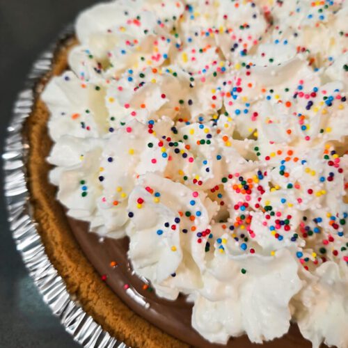 Close up photo of a chocolate cream pie in a graham cracker crust topped with whipped cream and rainbow sprinkles