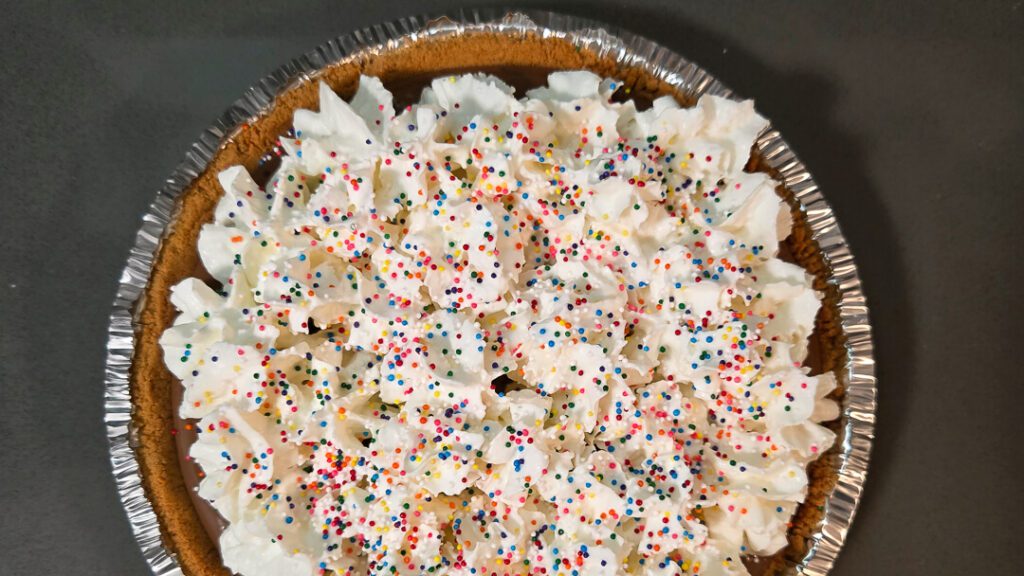 overhead photo of a chocolate cream pie decorated with rainbow sprinkles and whipped cream in a graham cracker crust