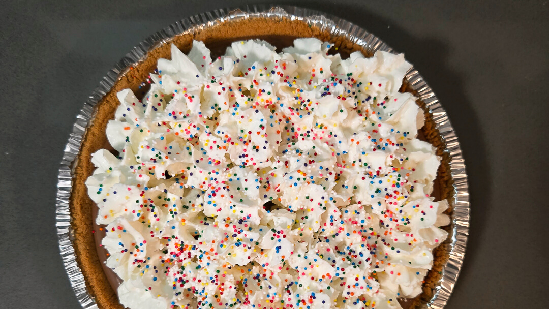 overhead photo of a chocolate cream pie decorated with rainbow sprinkles and whipped cream in a graham cracker crust