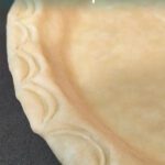 Partial close-up of a decorative unbaked pie crust with pressed arc-shaped pattern on the rim.