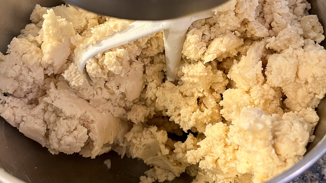 Close-up of pie dough being mixed in a stand mixer bowl with a paddle attachment, showing crumbly texture.