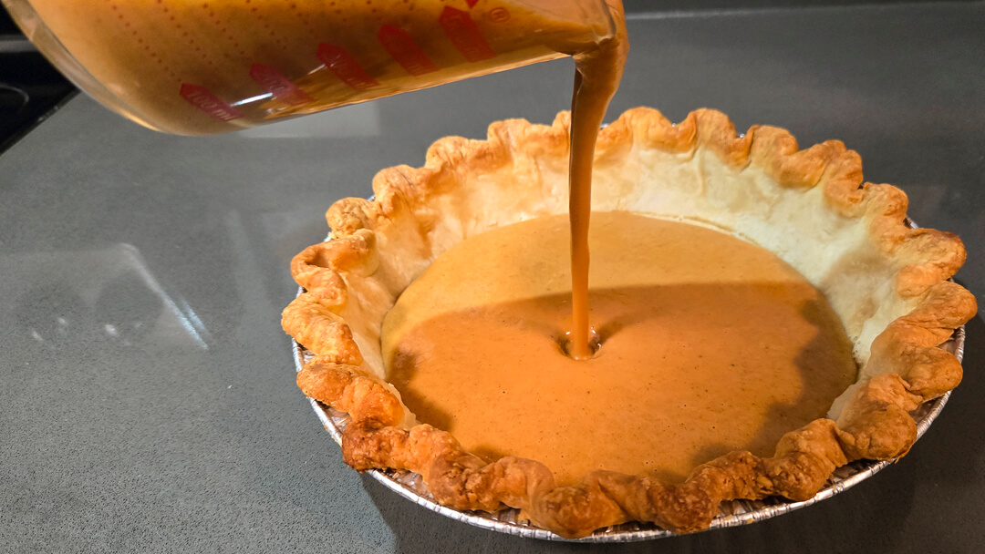 Pumpkin pie filling being poured from a measuring cup into a baked pie crust on a gray countertop.