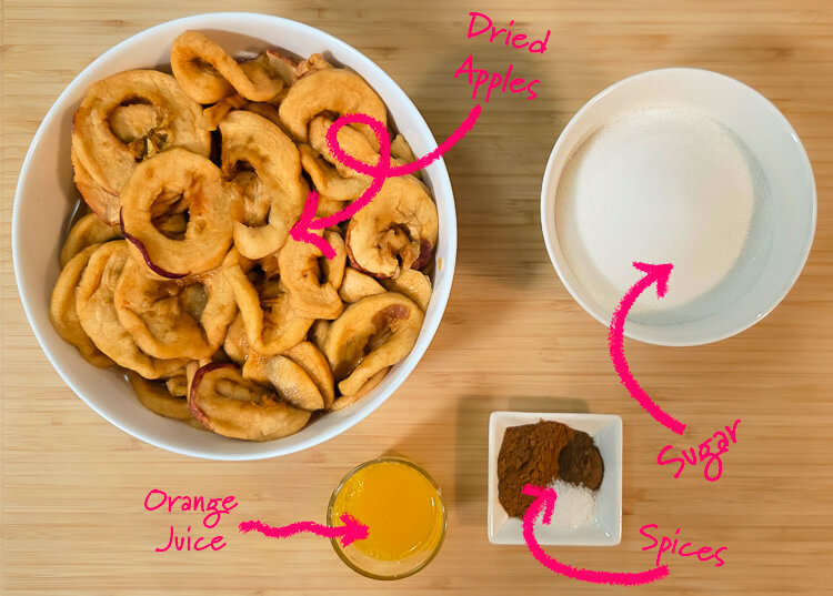 Ingredients for schnitz pie filling laid out on a cutting board with text labels: dried apples, sugar, orange juice, and spices