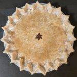 an overhead photo of a double crusted pie before it is baked. It is crimped with pointed flutes, has a star cutout in the center, and is brushed with cream and topped with demerara sugar