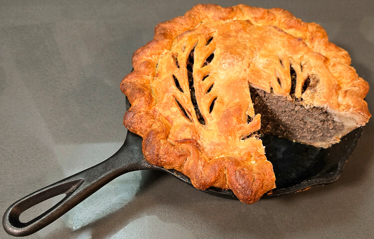 a photo of a tourtière baked in a cast iron pan with a slice cut out so you can see the meat filling inside