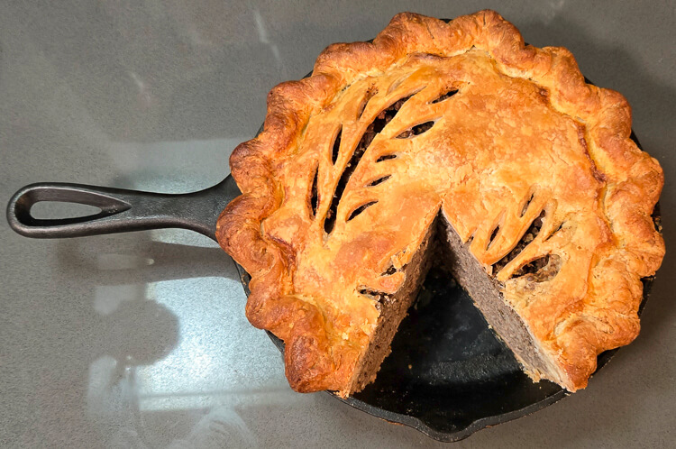 a horizontal photo of a baked tourtiere in a cast iron pan with a slice cut out