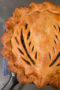 close up overhead photo of a half of a golden baked tourtière in a cast iron pan with wheat stalk vents cut into the crust