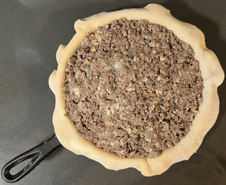 overhead photo of a tourtière being filled in a cast iron pan with the meat filling visible and no top crust