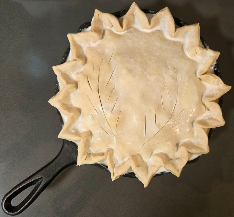 overhead photo of an unbaked tourtière in a cast iron pan with pointed flutes and decorative wheat stalk vents