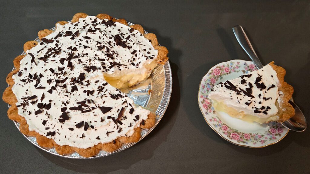 a photo of a banana cream pie topped with whipped cream and chocolate shavings with a slice cut out and placed next to it on a floral plate