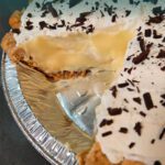 Slice of banana cream pie in a flaky crust, filled with homemade vanilla pudding and topped with whipped cream and chocolate shavings, shown with a missing slice in the pan.