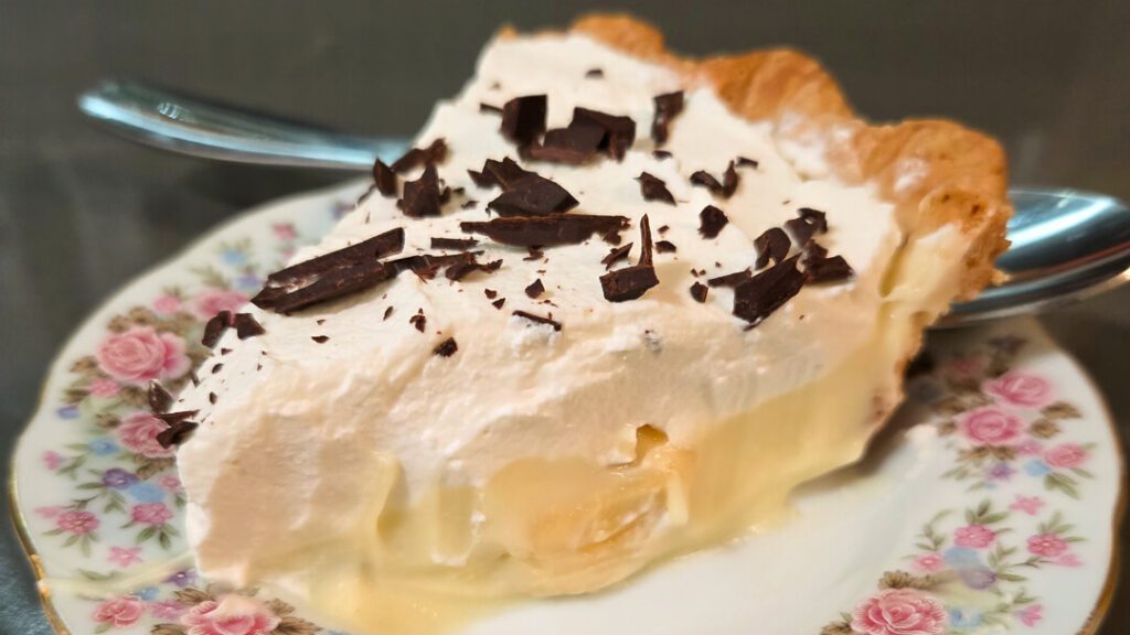 a close up photo of a slice of banana cream pie with homemade vanilla pudding, banana slices, topped with whipped cream and chocolate shavings in a flaky pie crust on an antique floral plate with roses
