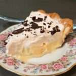 a close up photo of a slice of banana cream pie with homemade vanilla pudding, banana slices, topped with whipped cream and chocolate shavings in a flaky pie crust on an antique floral plate with roses