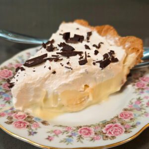 a close up photo of a slice of banana cream pie with homemade vanilla pudding, banana slices, topped with whipped cream and chocolate shavings in a flaky pie crust on an antique floral plate with roses