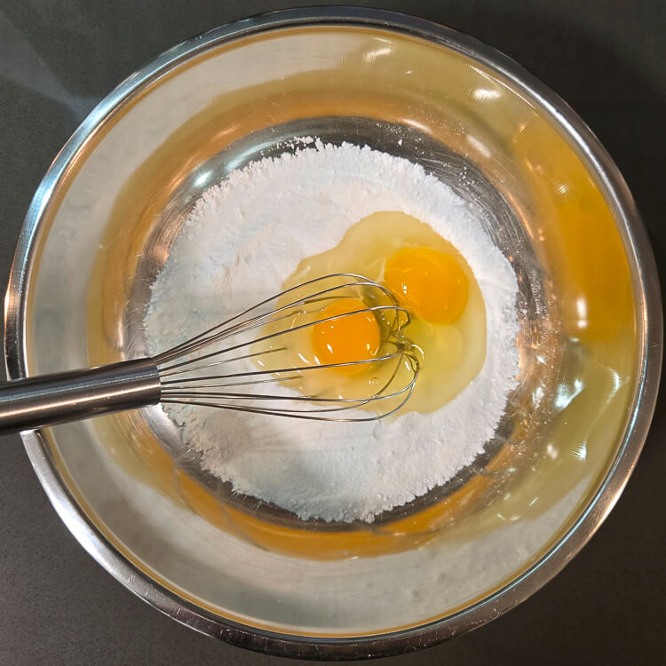 an overhead photo of eggs and flour in a metal mixing bowl with a balloon whisk