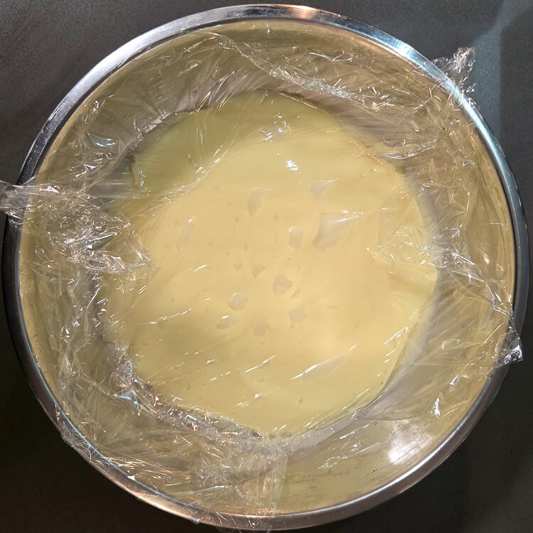 vanilla pudding in a mixing bowl with a piece of plastic pressed to the surface so it doesn't form a skin before it cools