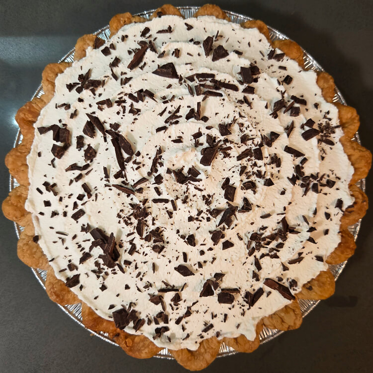 an overhead photo of a banana cream pie topped with whipped cream and chocolate shavings in a crimped pie crust