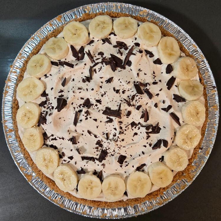 overhead photo of a completed banoffee pie with banana slices around the edge, and chocolate shavings sprinkled over the whipped cream