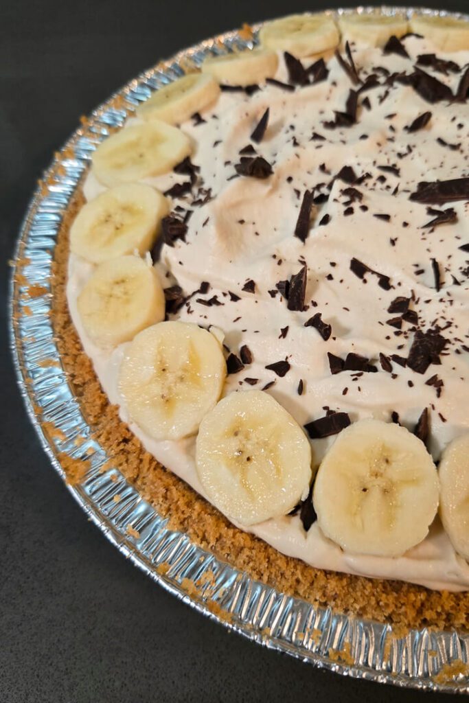 A close up photo of a banoffee pie with a golden brown crust made with digestive biscuits, banana slices around the edge, and chocolate shavings atop the coffee flavored whipped cream