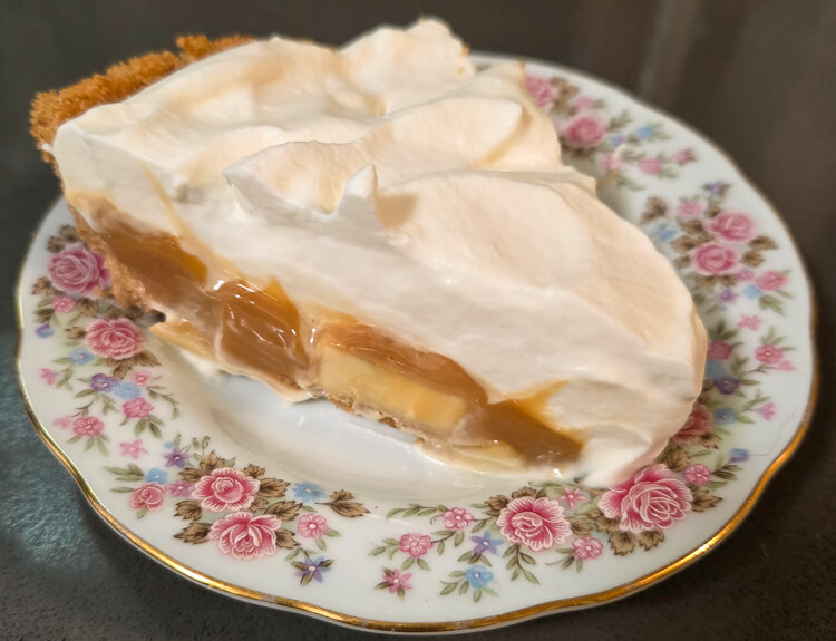 a close up photo of a slice of banoffee pie with banana slices, glossy dulce de leche caramel, and coffee whipped cream in a golden digestive biscuit crumb crust on a floral antique plate