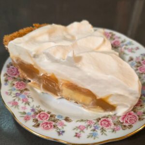 a close up photo of a slice of banoffee pie with banana slices, glossy dulce de leche caramel, and coffee whipped cream in a golden digestive biscuit crumb crust