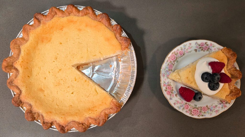 an overhead photo of a buttermilk pie with a slice cut out and placed next to it on a floral plate served with whipped cream and fresh berries