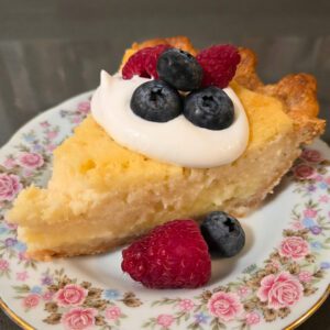 a close up photo of a slice of buttermilk pie on a floral antique plate served with a dollop of whipped cream and fresh blueberries and raspberries