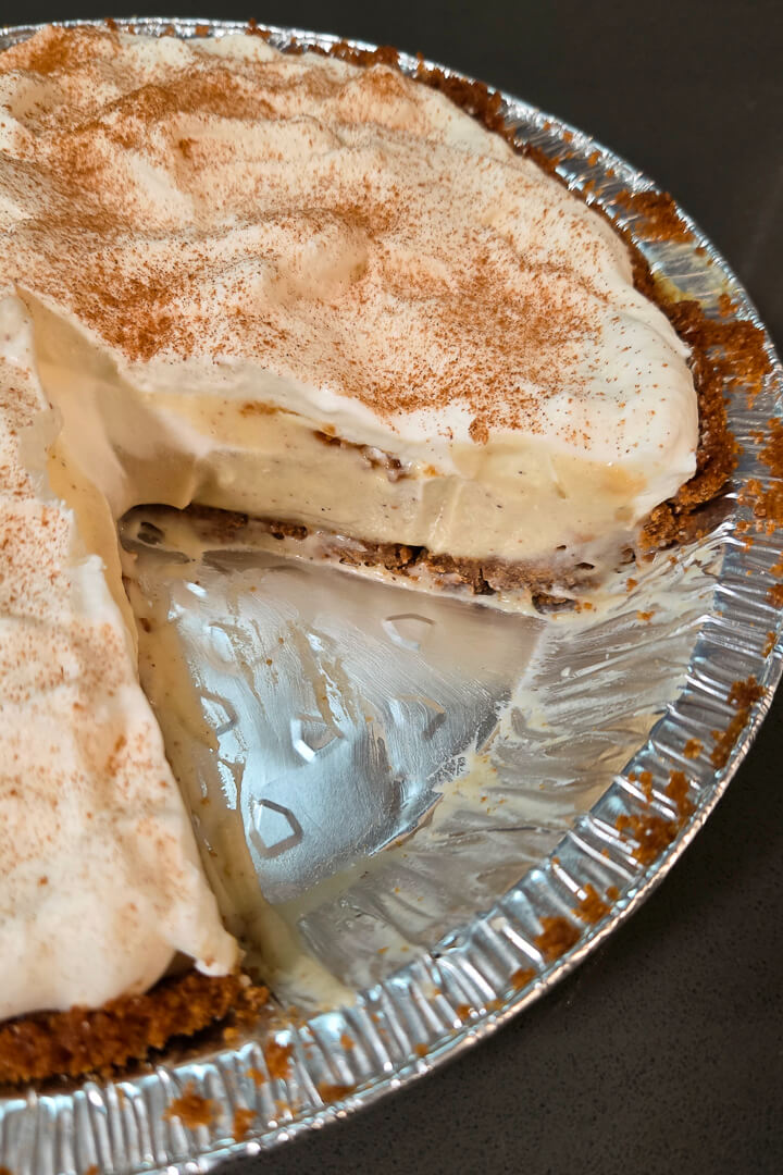 close up photo of an eggnog cream pie with a slice cut out showing homemade eggnog pudding in a gingersnap pie crust with a whipped cream topping dusted with cinnamon