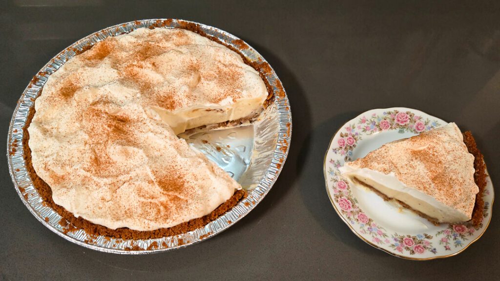 overhead photo of an eggnog cream pie with homemade eggnog pudding, with a slice cut out and placed on an antique floral plate next to it