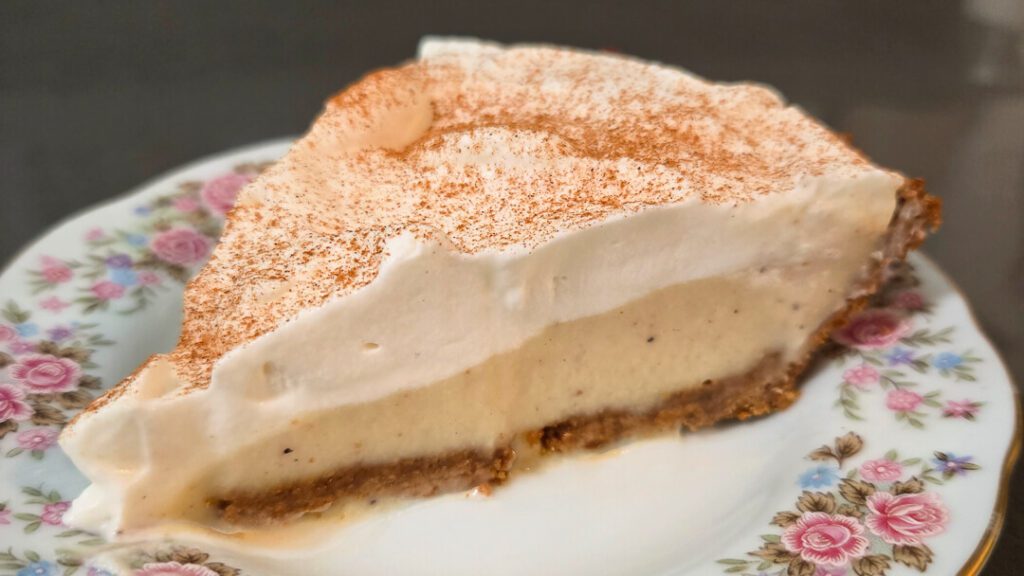 a slice of eggnog cream pie with eggnog pudding underneath a layer of whipped cream in a gingersnap crust on an antique floral plate dusted with cinnamon