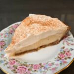 a slice of eggnog cream pie with eggnog pudding underneath a layer of whipped cream in a gingersnap crust on an antique floral plate