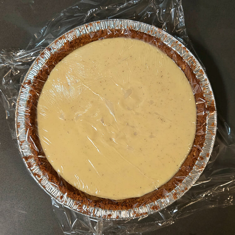 overhead photo of an eggnog cream pie filled with freshly made warm eggnog pudding with a piece of plastic wrap laid over top so it doesn't form a skin