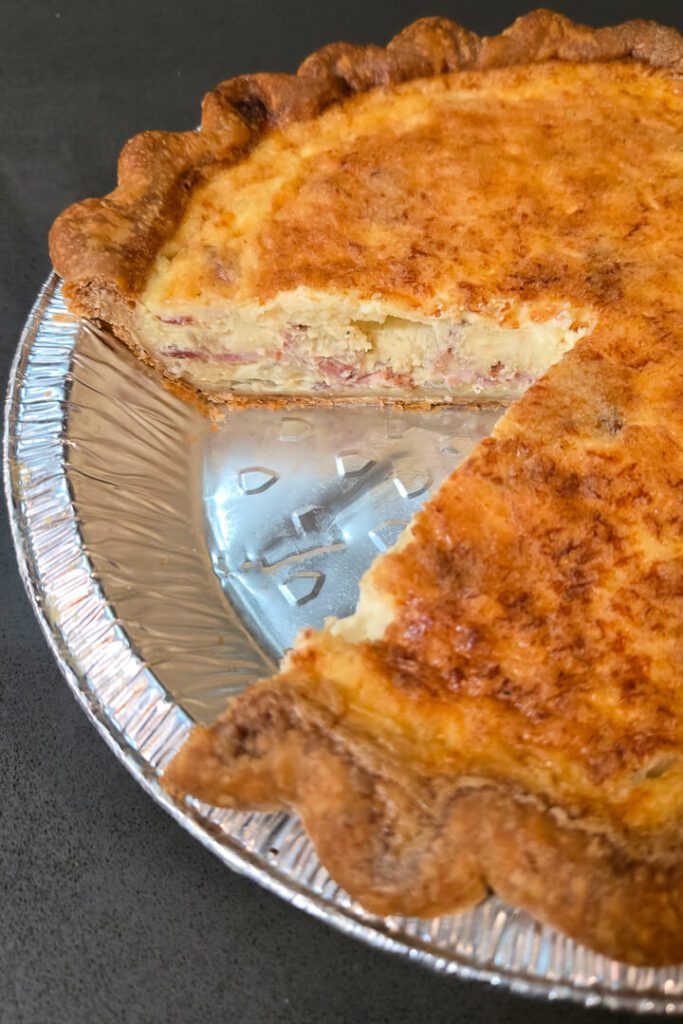 a close up photo of a quiche lorraine with a slice cut out so you can see the cross section with flaky pie crust, bacon, egg custard, and onions